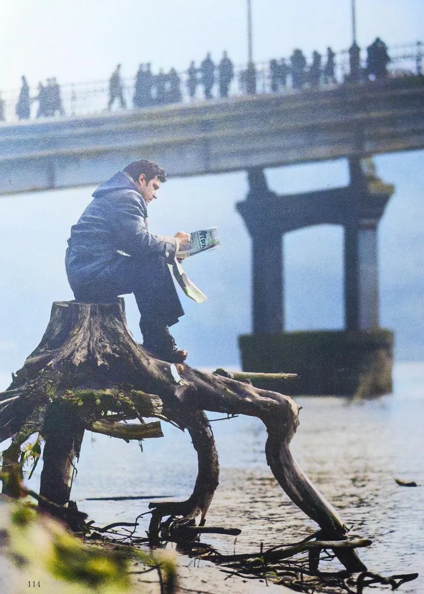 A man is reading something while sitting on a stump on the bank of the Dnieper River with a view of the bridge